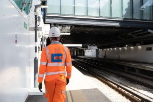 A person walking along a train station platform wearing orange Costain branded personal protective equipment