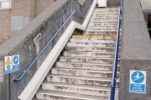 A screenshot of part of a video of Cardonald station showing accessibility improvements to the stairs leading to the station, which appear to have new blue handrails and yellow and white painted stairs