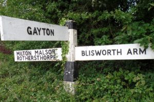 A stock photo of an old wooden signpost with one arm pointing towards GAYTON and the sign is nestled into a hedgerow or bush